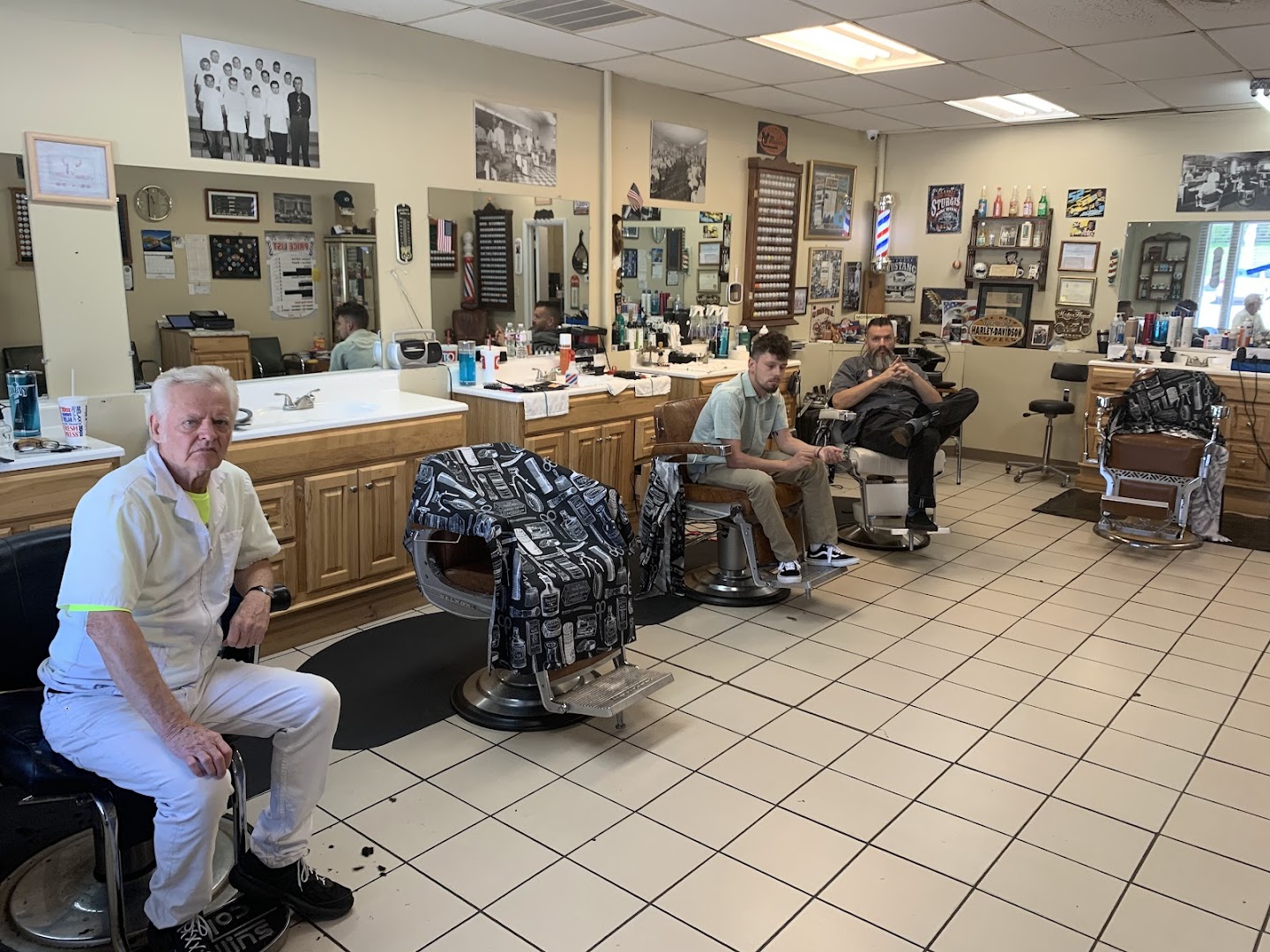 Inside Gills Barbershop — three chairs, mirrors, a vintage class photo of barbers on the back wall, a US flag, an older client sitting in the foreground.